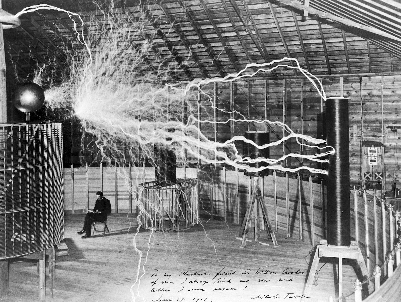 Nikola Tesla seated calmly beside his giant coil as long electric arcs crackle overhead, a double-exposed photograph from his Colorado Springs laboratory